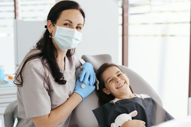 Uma dentista sentada ao lado de uma criança sorrindo sentada em uma cadeira odontológica