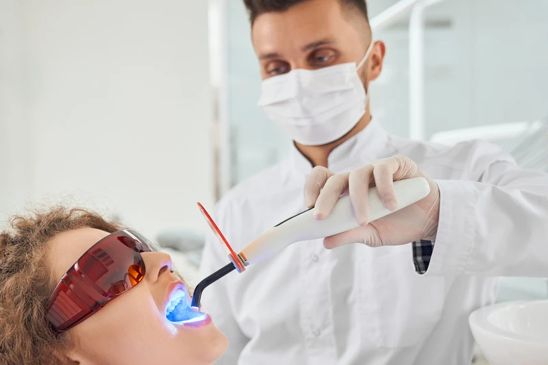 Dentista realizando um procedimento de clareamento dental em uma paciente com óculos de proteção, iluminando a boca com uma luz azul.