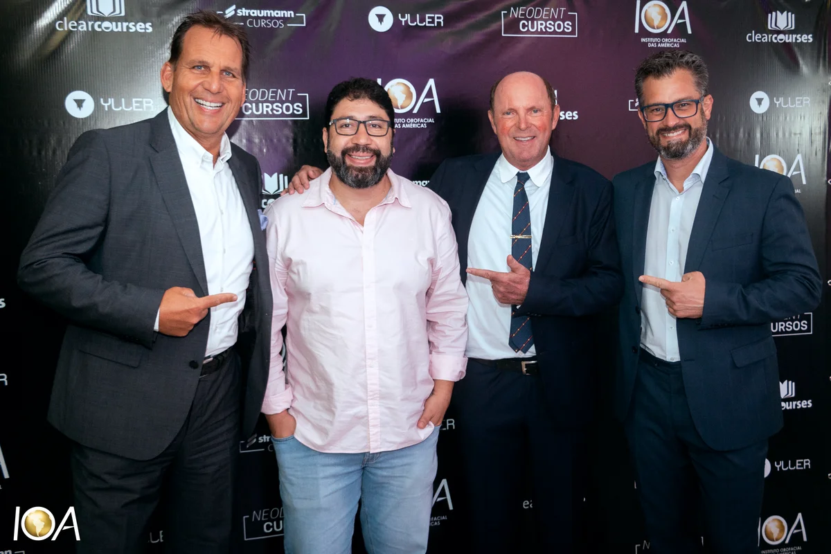 Quatro homens sorrindo em evento corporativo com fundo de patrocinadores, três deles apontando para o homem de camisa rosa no centro.
