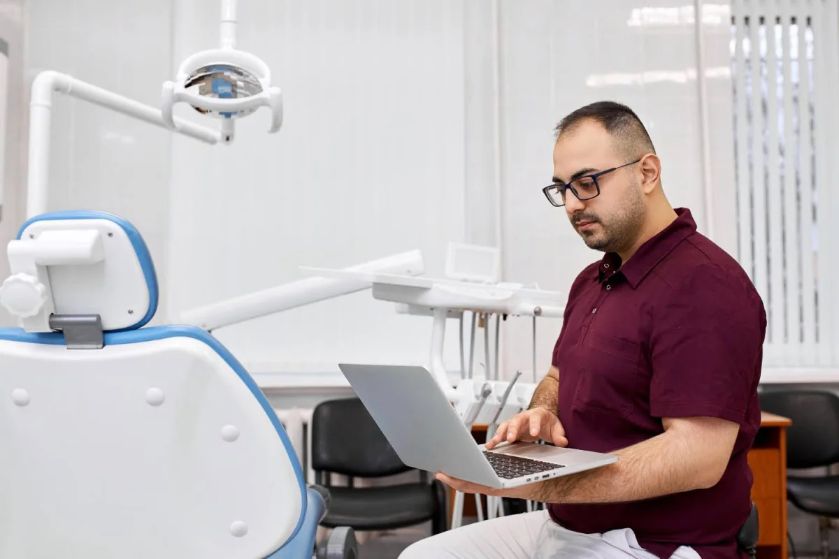 Dentista usando laptop em consultório odontológico moderno com cadeira odontológica azul e equipamentos ao fundo